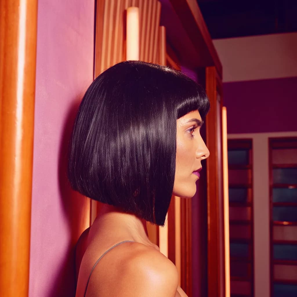 Colombian woman with a sleek shiny bob haircut, side profile, glossy dark hair, pink backdrop, studio-quality daylight